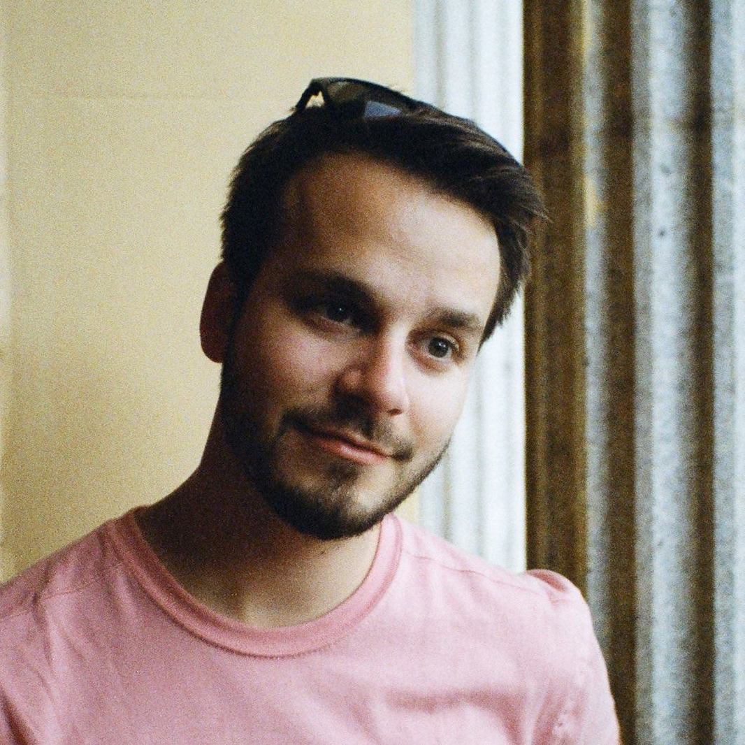 Matt Ahl headshot wearing pink t-shirt, with dark hair and short beard and glasses atop his head